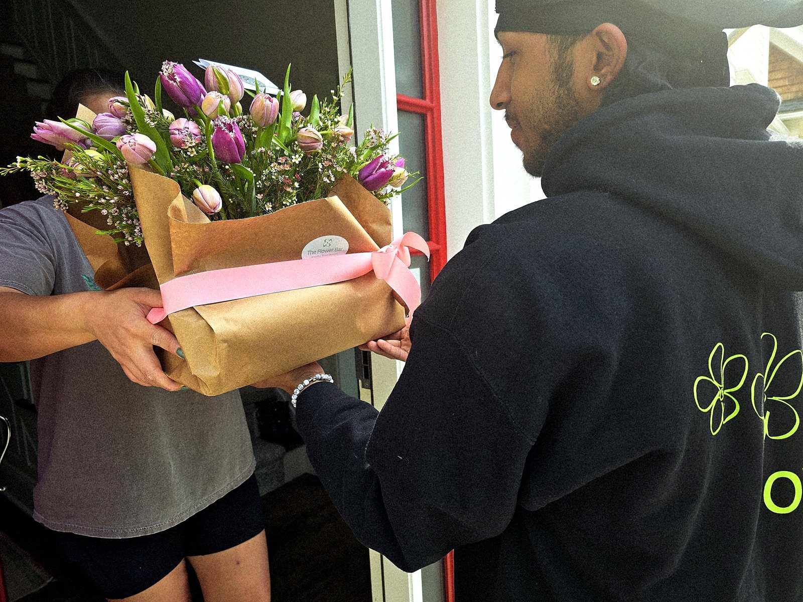 A Flower Bar delivery driver handing a kraft-wrapped bouquet of purple tulips to a recipient at her front door in Westchester, NY
