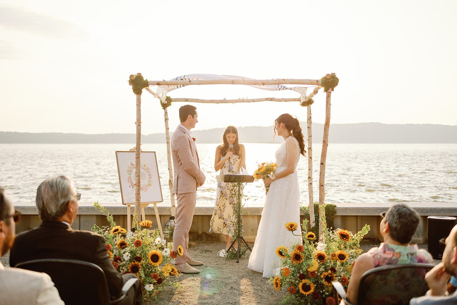 Waterfront summer wedding ceremony in Westchester beneath a birch chuppah, with sunflower and wildflower arrangements by The Flower Bar