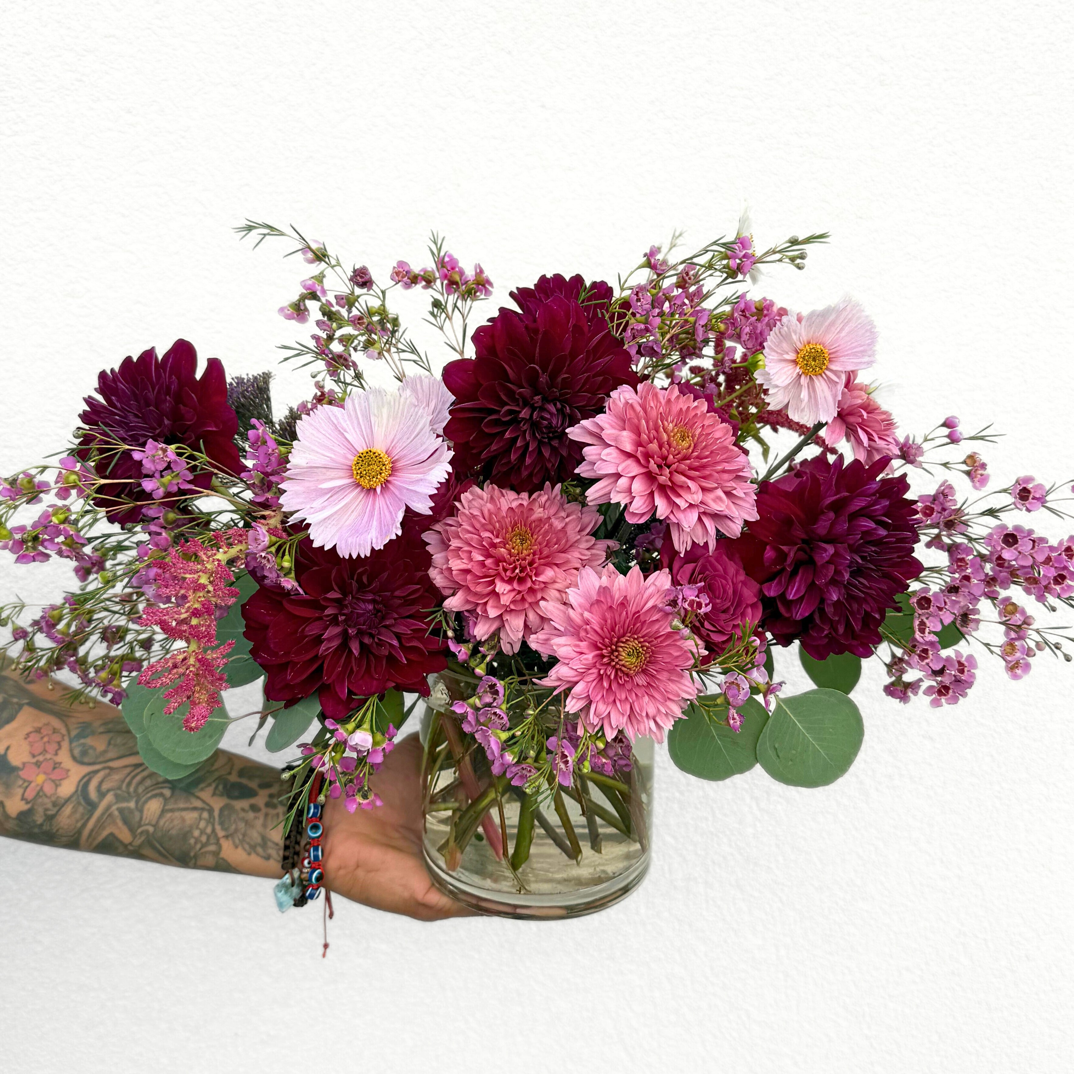 A hand holding a clear glass vase filled with deep burgundy and pink dahlias, light pink gerberas, and purple waxflower stems against a white background.