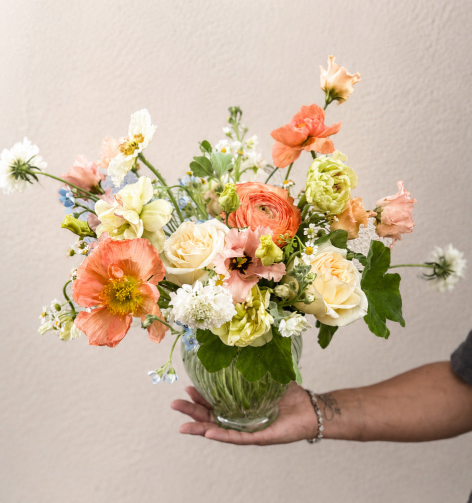 Hand holding a clear glass vase filled with coral ranunculus, cream roses, white astilbe, and greenery in a lush, rounded arrangement.