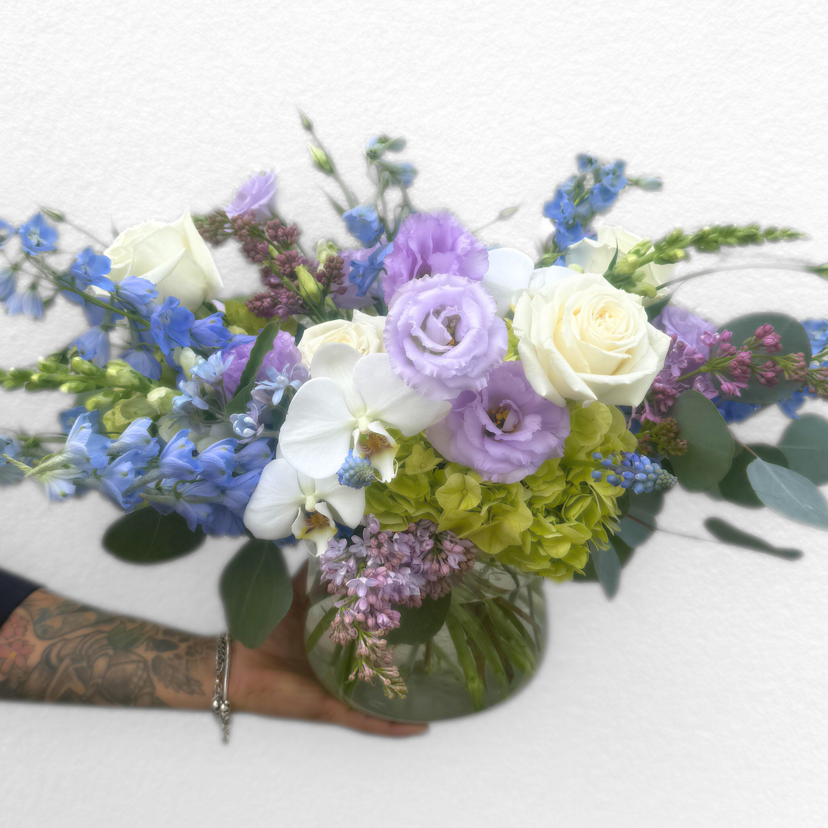 Lavender roses, white orchids, blue delphinium, and green hydrangea arranged in a clear glass vase with lush greenery.
