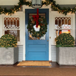 Blue front door decorated with a lush green wreath adorned with a red velvet bow, flanked by golden topiary topiaries in concrete planters.