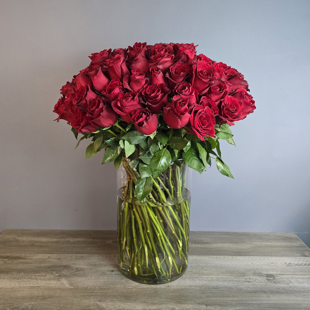 A lush bouquet of deep red roses arranged in a clear glass cylinder vase with green stems and foliage visible in water.