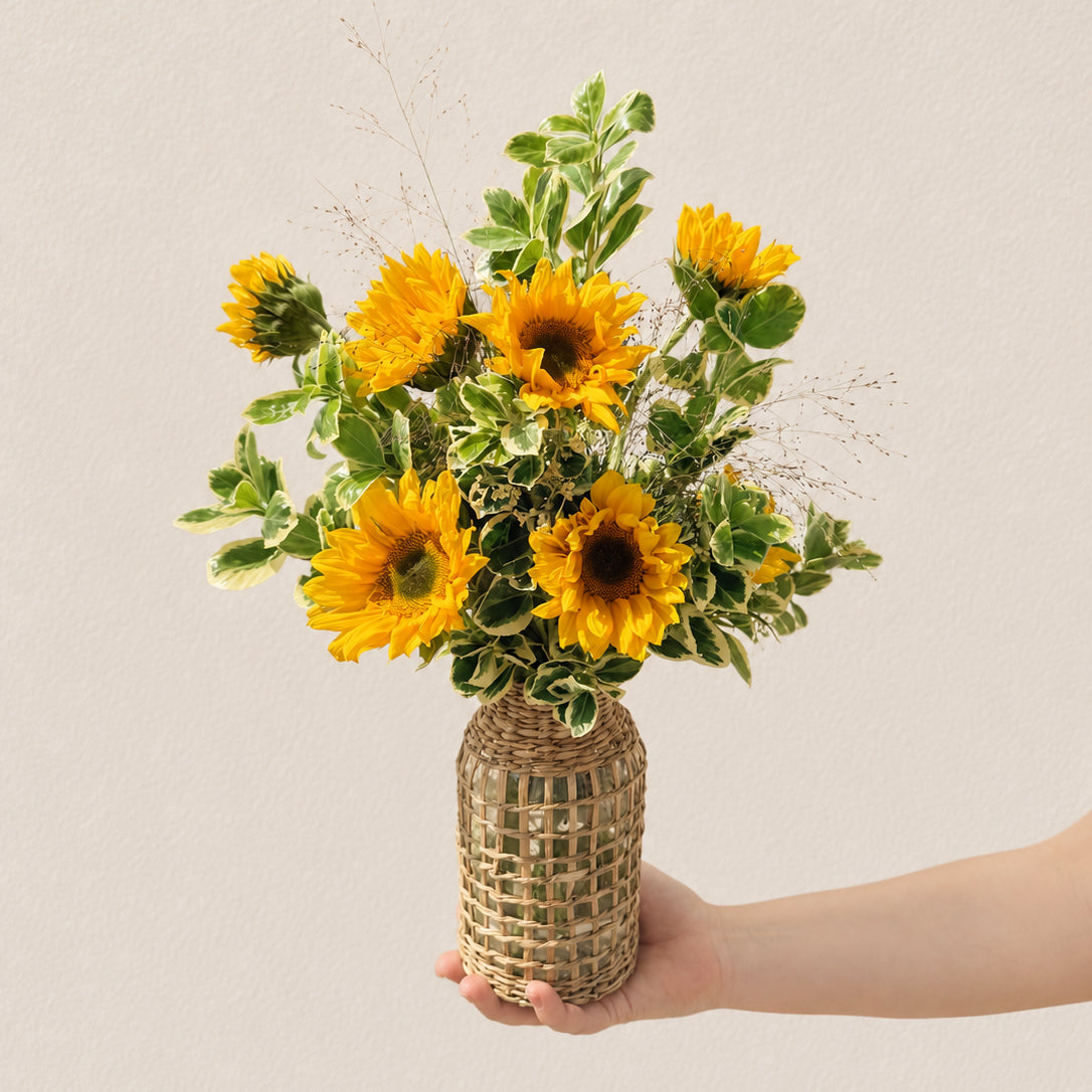 Signature sunflower arrangement with ten sunflowers and lush greens in a tall wicker bottle-neck vase