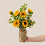 Signature sunflower arrangement with ten sunflowers and lush greens in a tall wicker bottle-neck vase
