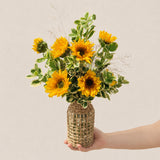 Signature sunflower arrangement with ten sunflowers and lush greens in a tall wicker bottle-neck vase