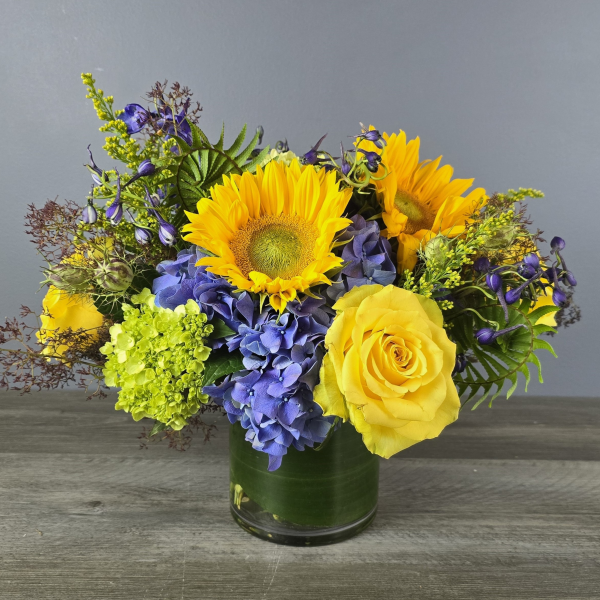 Vibrant mixed bouquet featuring sunflowers, yellow roses, blue hydrangeas, and green chrysanthemums in a dark green cylinder vase.