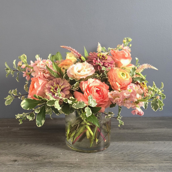 Lush garden-style arrangement featuring coral roses, peach ranunculus, burgundy dahlias, and green foliage in a clear glass vase.