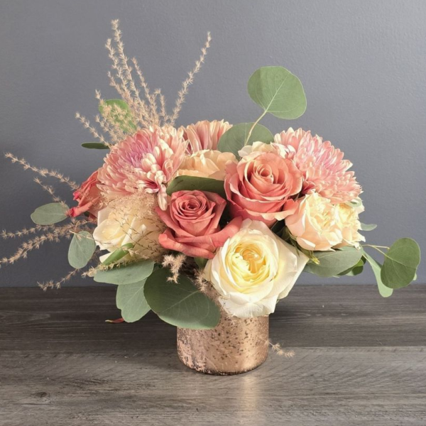 A textured cork vase holds pink roses, cream dahlias, yellow garden roses, and dried grasses with eucalyptus greenery.