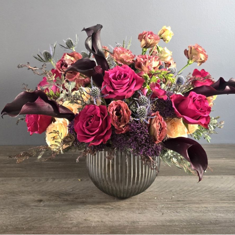 Luxe floral arrangement featuring deep magenta roses, burgundy calla lilies, coral carnations, and cream roses in a ribbed silver vase. — The Flower Bar, Larchmont NY