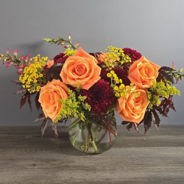 A glass vase holds a lush arrangement of coral roses, deep burgundy dahlias, and yellow solidago with cascading foliage.