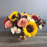 Vibrant arrangement of sunflowers, roses in peach and burgundy, dahlias, and white flowers in a bronze pedestal vase with dark foliage.
