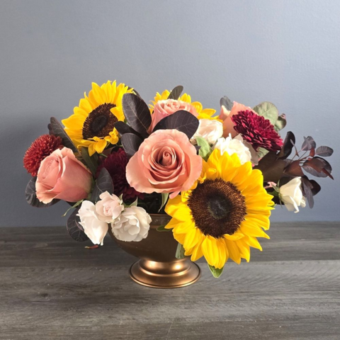 Vibrant arrangement of sunflowers, roses in peach and burgundy, dahlias, and white flowers in a bronze pedestal vase with dark foliage. — The Flower Bar, Larchmont NY