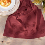 Burgundy linen napkin embroidered with autumn leaves arranged on a wooden table next to a plate of sliced apples.