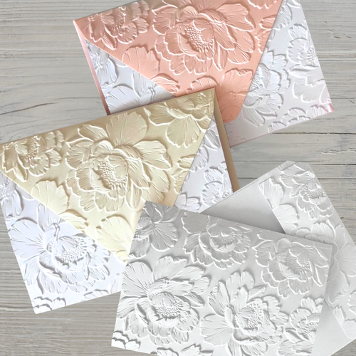 Three embossed floral greeting cards in white, cream, and coral featuring detailed peony designs arranged on a wooden surface.