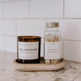 A minimalist marble tray displays a "Relaxation" scented candle in dark glass and a glass jar of safety matches against white subway tile.