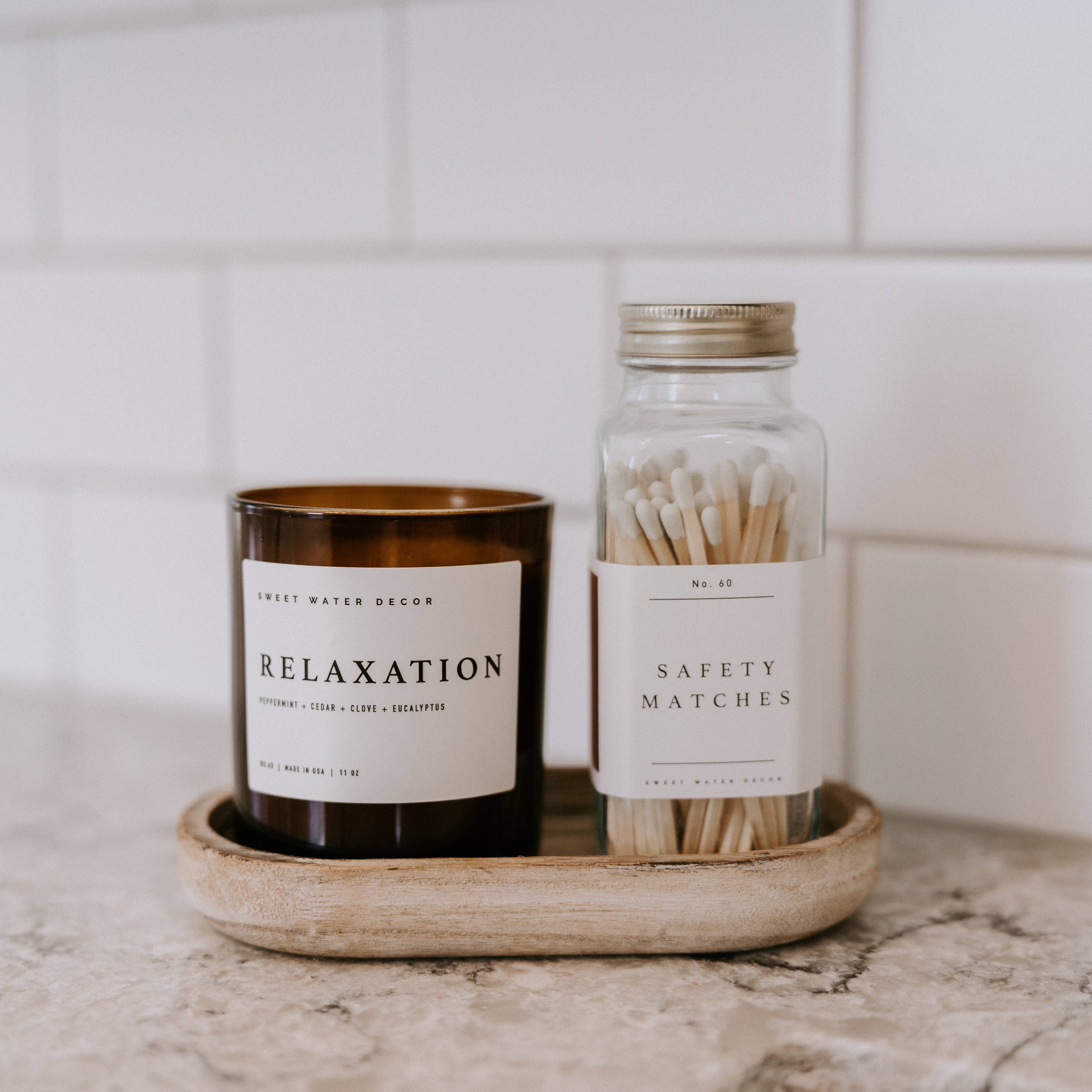A minimalist marble tray displays a "Relaxation" scented candle in dark glass and a glass jar of safety matches against white subway tile.