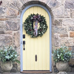 Cream-colored arched door adorned with a lush evergreen wreath tied with a purple plaid ribbon, flanked by potted eucalyptus arrangements.