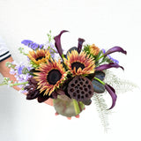 A vibrant hand-tied bouquet featuring golden sunflowers, purple limonium, deep burgundy calla lilies, and delicate fern accents in a clear glass vase.