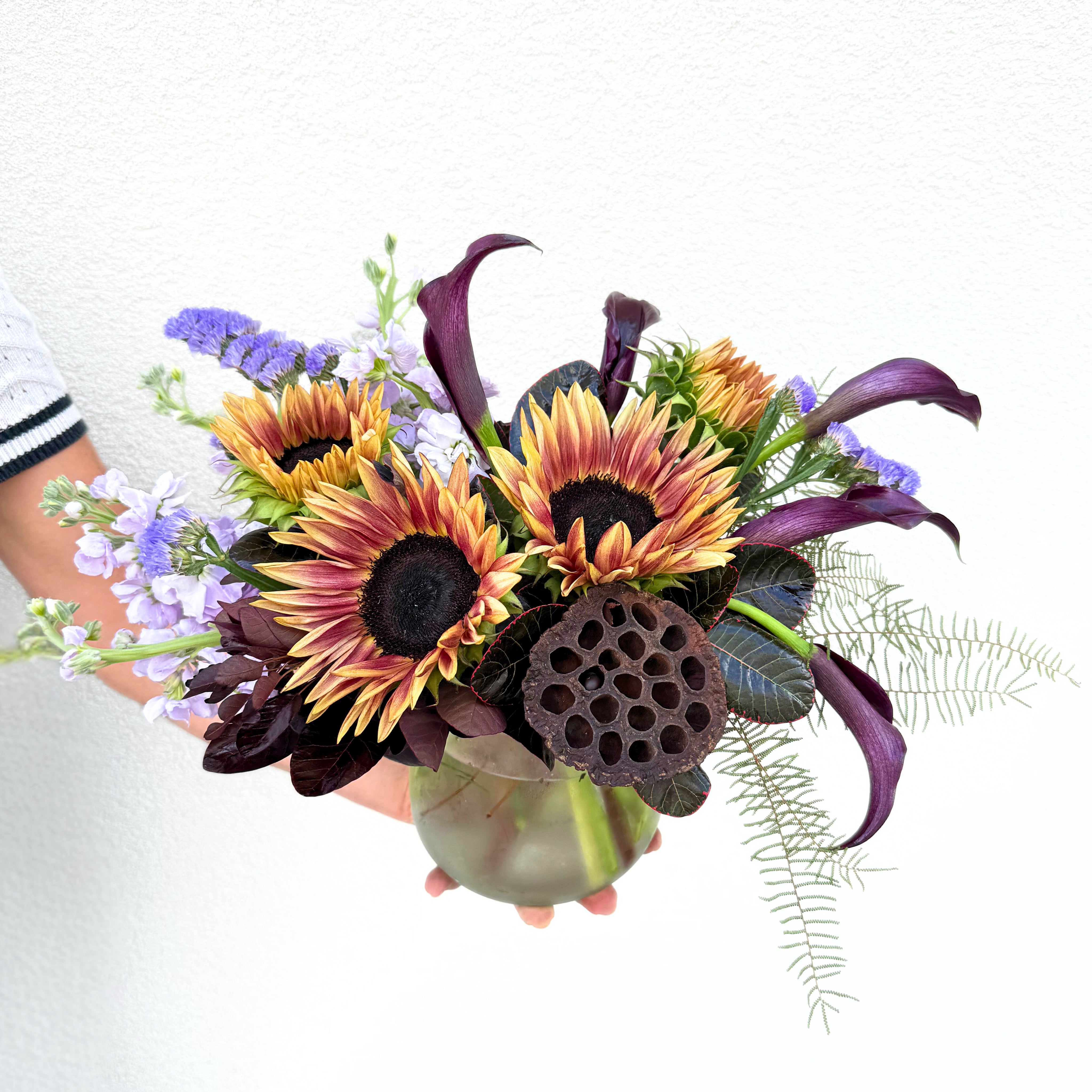 A vibrant hand-tied bouquet featuring golden sunflowers, purple limonium, deep burgundy calla lilies, and delicate fern accents in a clear glass vase.
