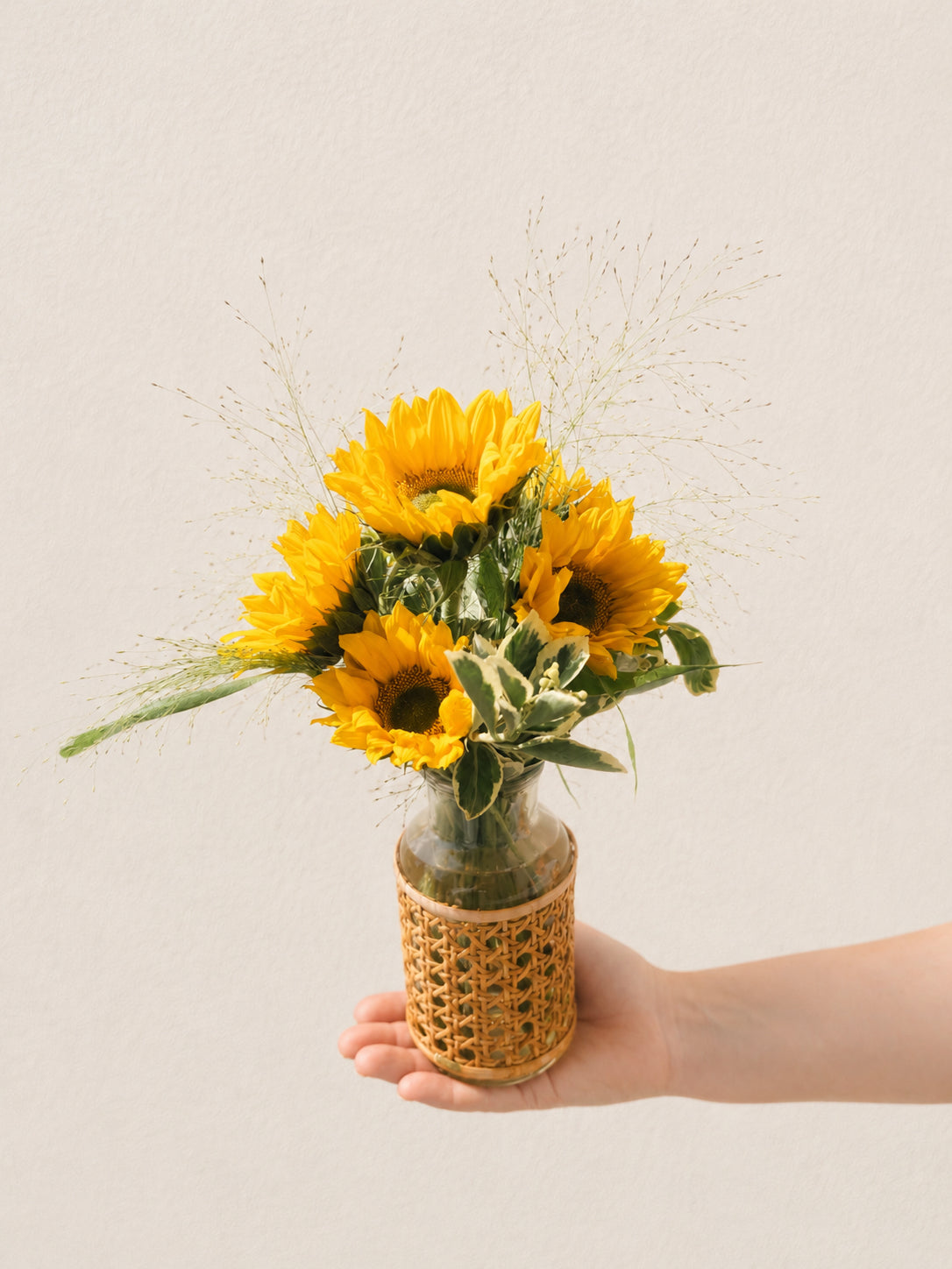 Mini Soleil sunflower arrangement with five golden sunflowers and airy grasses in a small wicker bottle-neck vase