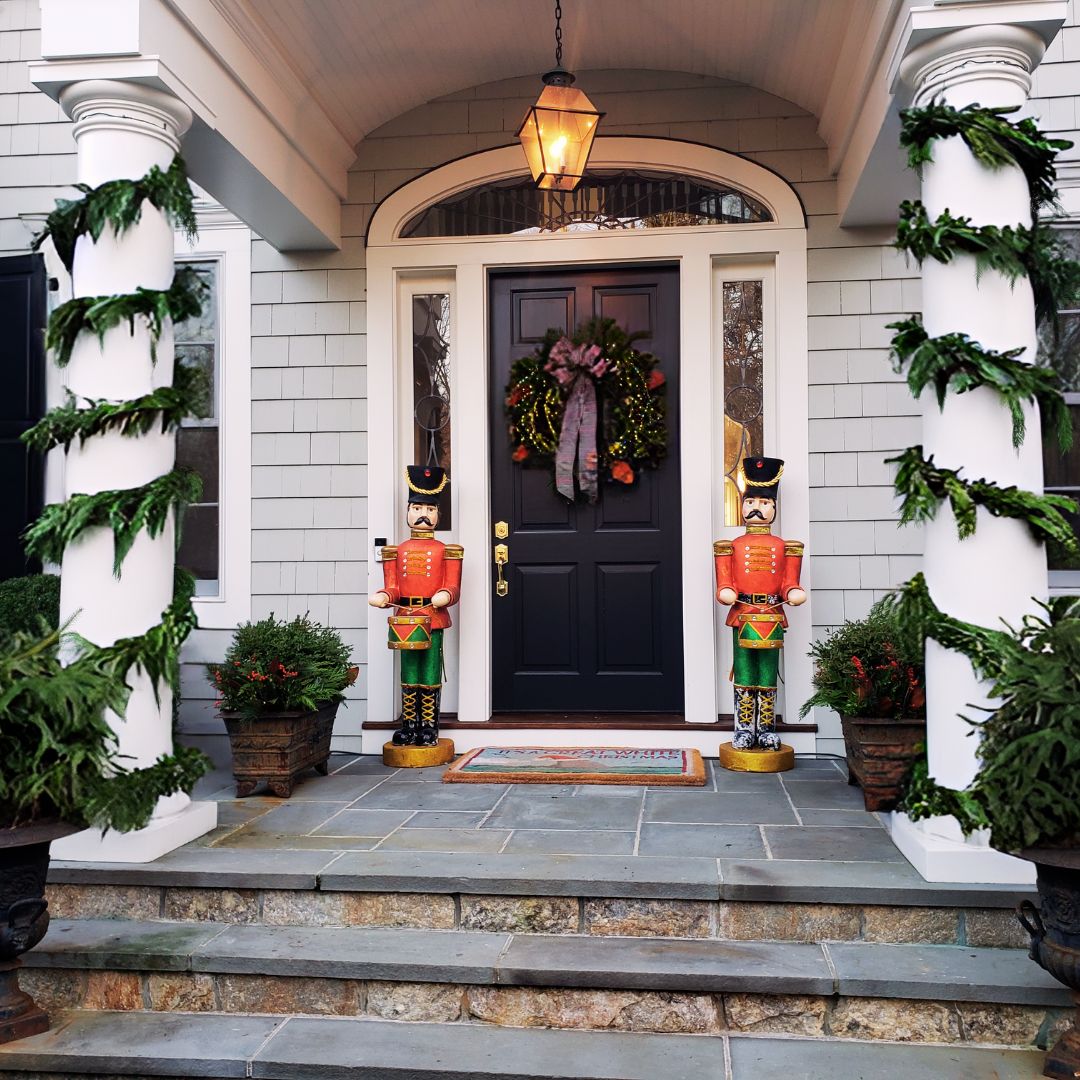 Elegant front entrance decorated with cascading garland swags, festive nutcracker statues, and poinsettia planters flanking a black door.