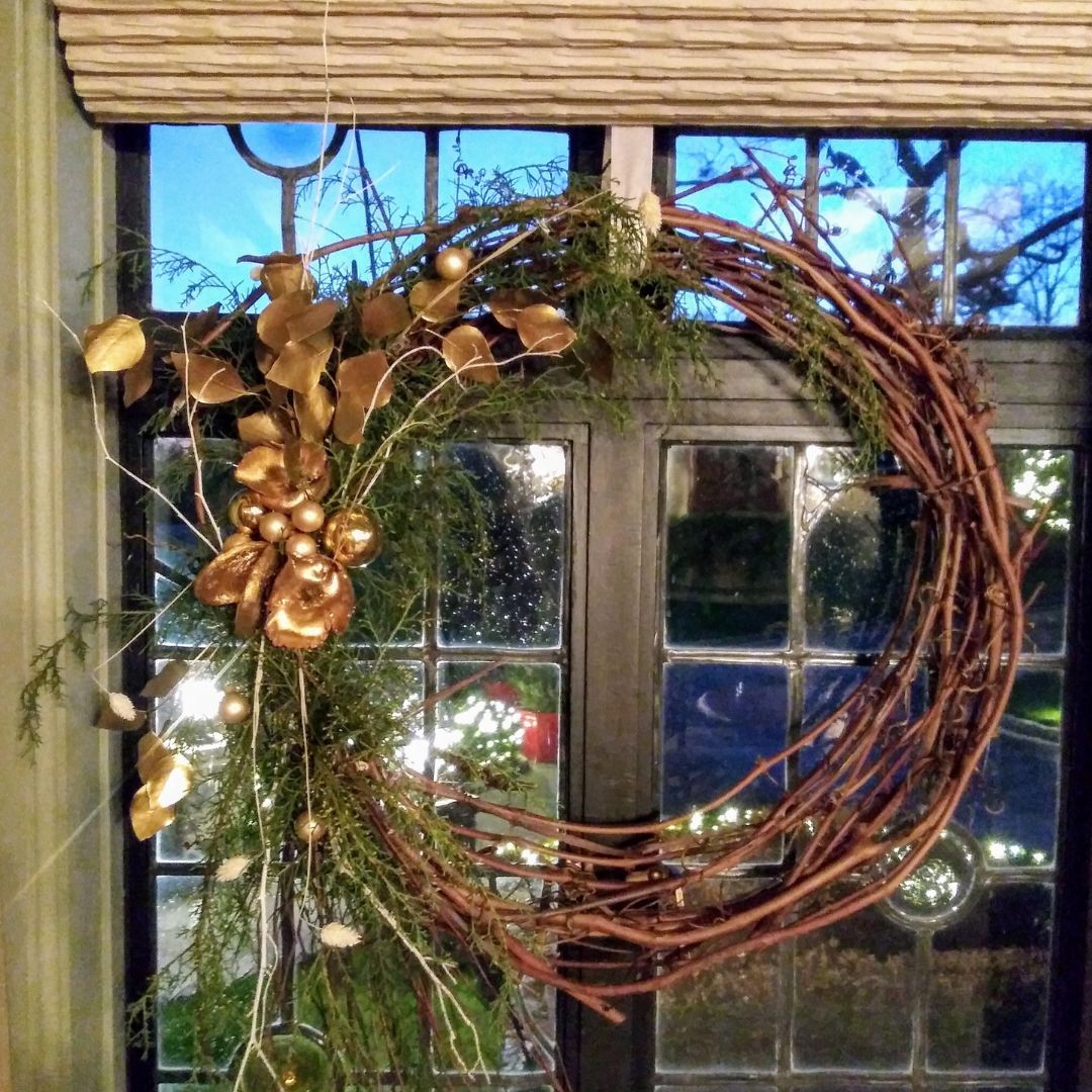 Rustic grapevine wreath adorned with golden leaves and fresh green foliage hanging on a glass door.