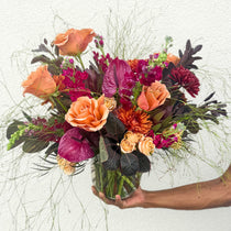A hand holds a vibrant vase arrangement of coral roses, burgundy dahlias, and dark purple orchids with wispy green stems and foliage.