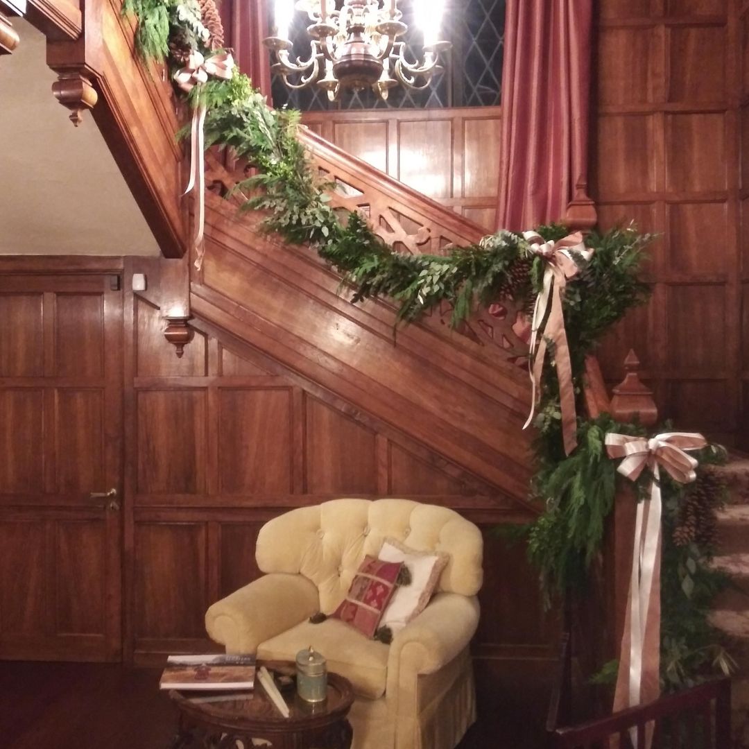 A cream-colored armchair sits beneath a wooden staircase decorated with a lush garland of greenery and cream-colored ribbon bows.