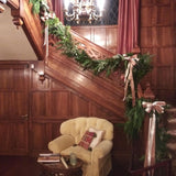 A cream-colored armchair sits beneath a wooden staircase decorated with a lush garland of greenery and cream-colored ribbon bows.