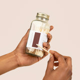 A person's hands holding a clear glass bottle with a white label containing pale yellow liquid against a beige background.