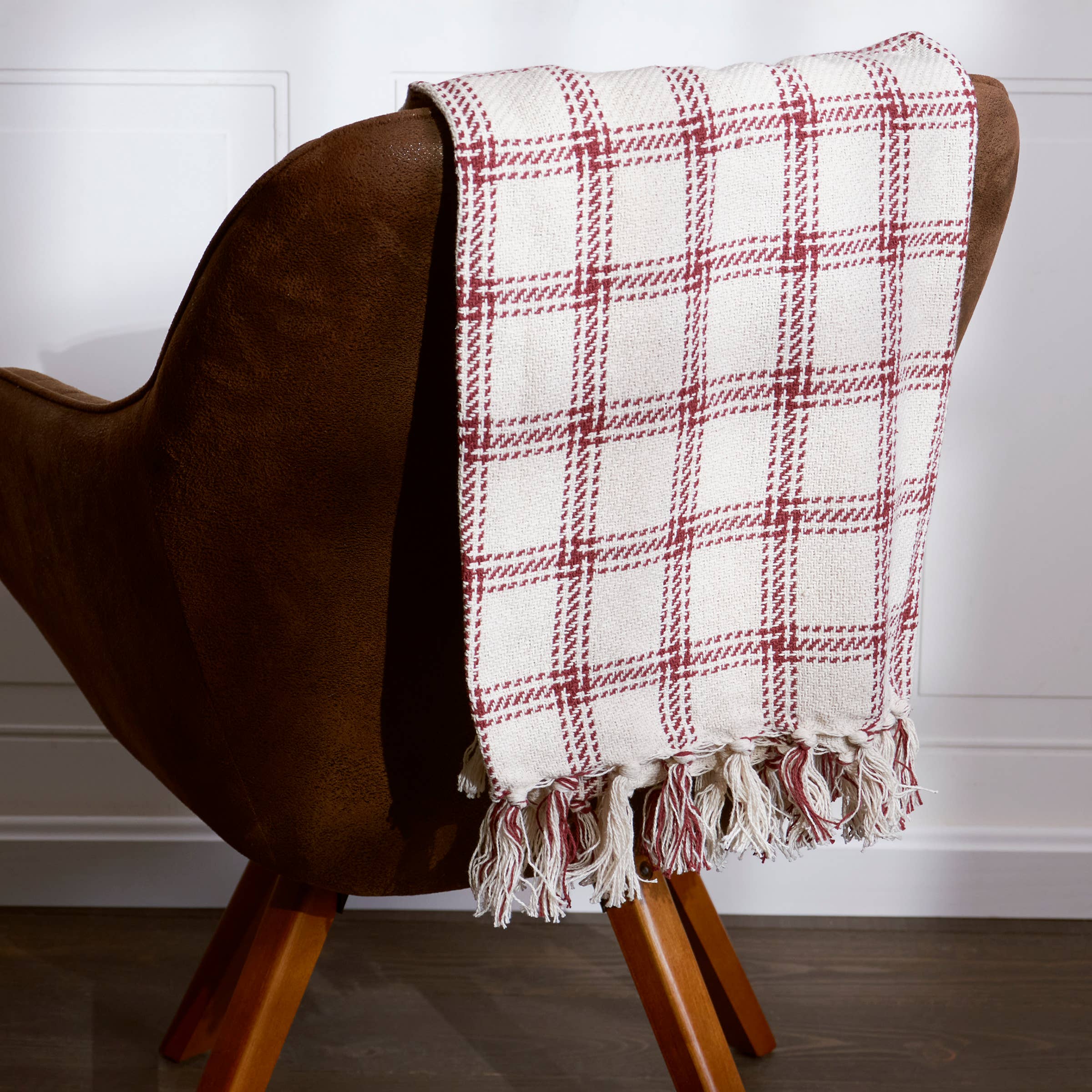 A red and cream plaid throw blanket drapes over a brown mid-century modern chair with wooden legs.