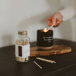A hand lights a black scented candle next to a glass jar of wooden matches on a dark surface.
