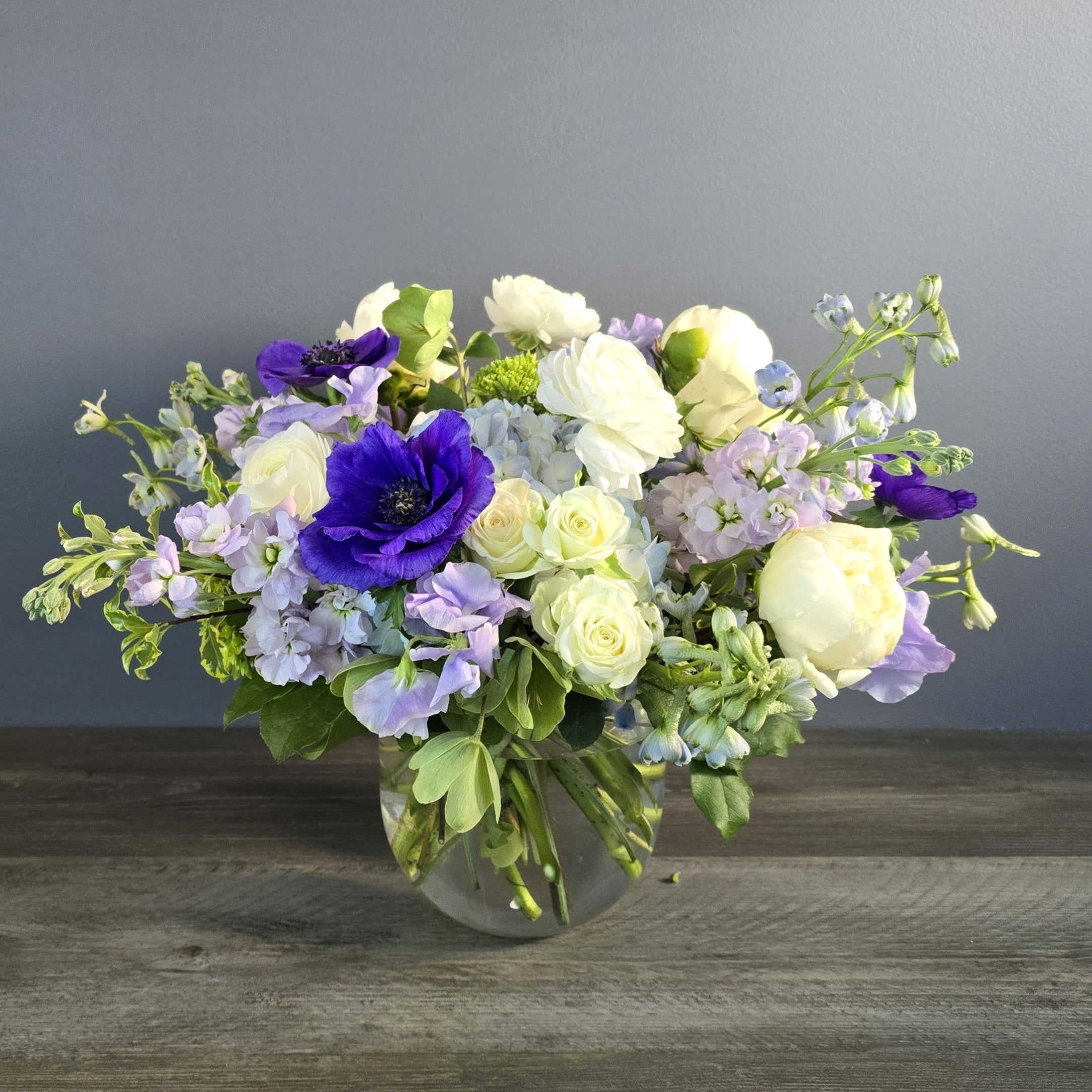 Lush garden-style arrangement in clear vase featuring purple anemones, white roses, lavender sweet peas, and chartreuse spray roses.
