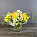 Bright yellow roses and white daisies arranged in a clear glass vase with green fern accents on a dark wooden surface.