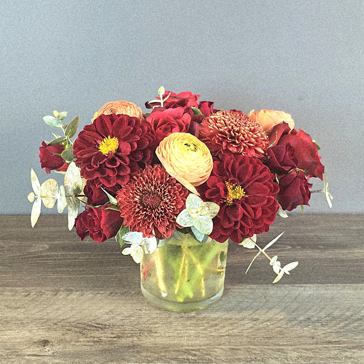 Deep red dahlias and ranunculus with cream-colored blooms and white jasmine arranged in a clear glass vase with green stems visible.