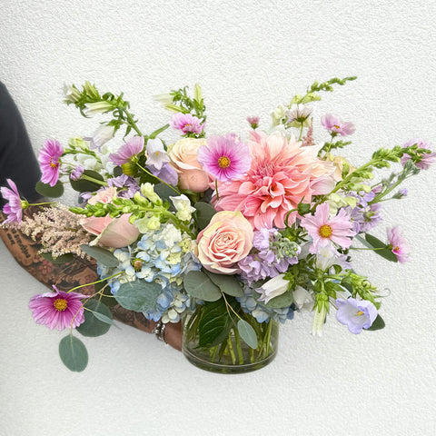 Pink dahlias, roses, and magenta gerbera daisies arranged in a clear glass vase with blue hydrangeas and purple lisianthus. — The Flower Bar, Larchmont NY