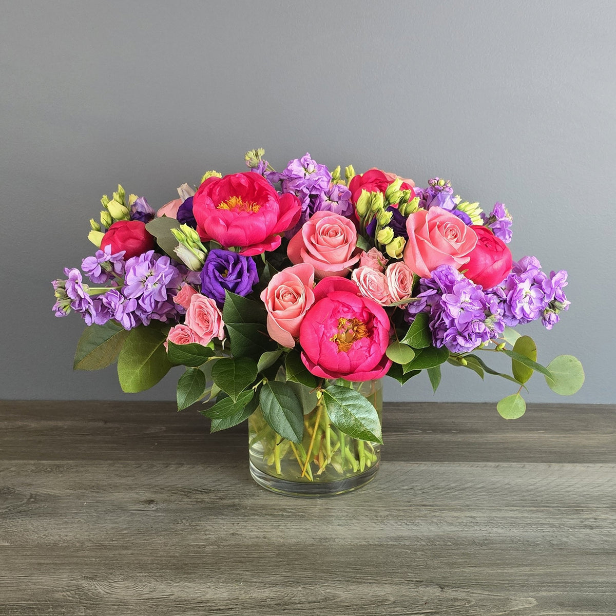 Vibrant mixed bouquet of hot pink peonies, purple lisianthus, coral roses, and green trick dianthus in a clear glass vase.
