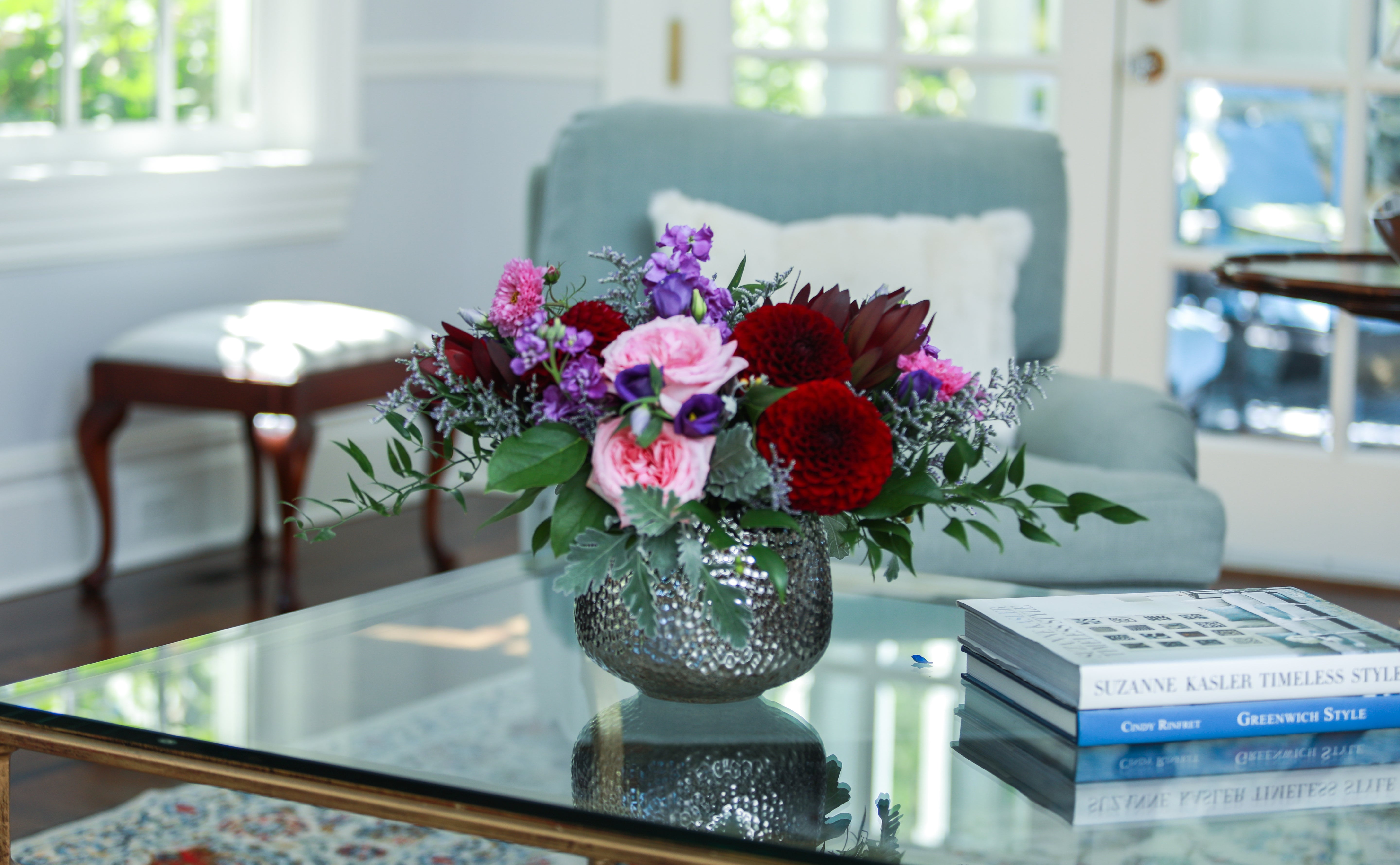 A vibrant floral arrangement of deep red dahlias, pink peonies, purple statice, and burgundy roses in a textured grey ceramic vase sits on a glass table.