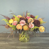 A lush mixed bouquet in a clear glass vase features pink roses, yellow ranunculus, burgundy dahlias, green trick dianthus, and dried grasses.
