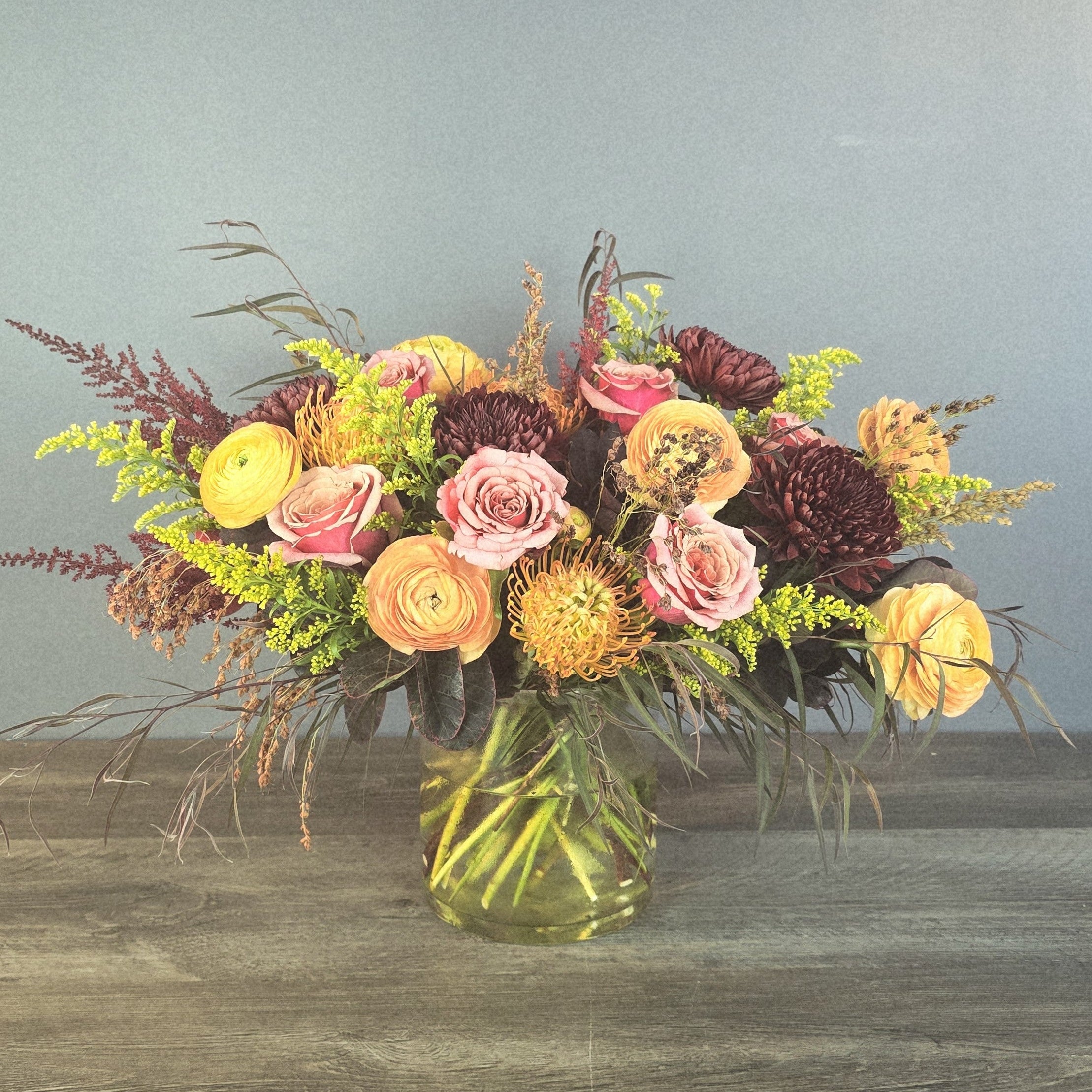 A lush mixed bouquet in a clear glass vase features pink roses, yellow ranunculus, burgundy dahlias, green trick dianthus, and dried grasses.