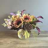 A vibrant autumn arrangement of red and yellow gerbera daisies, purple statice, burgundy calla lilies, and fern greenery in a clear glass vase.