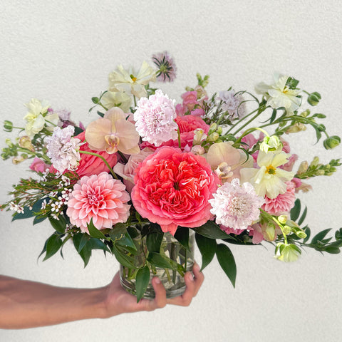A hand holding a glass vase with a lush garden-style arrangement of coral roses, pink dahlias, peach orchids, and cream daisies with greenery. — The Flower Bar, Larchmont NY