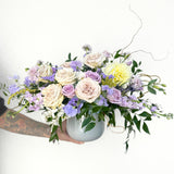 Lush mixed bouquet in white ceramic vase featuring cream roses, lavender and pink spray roses, purple lisianthus, yellow dahlia, and cascading greenery.