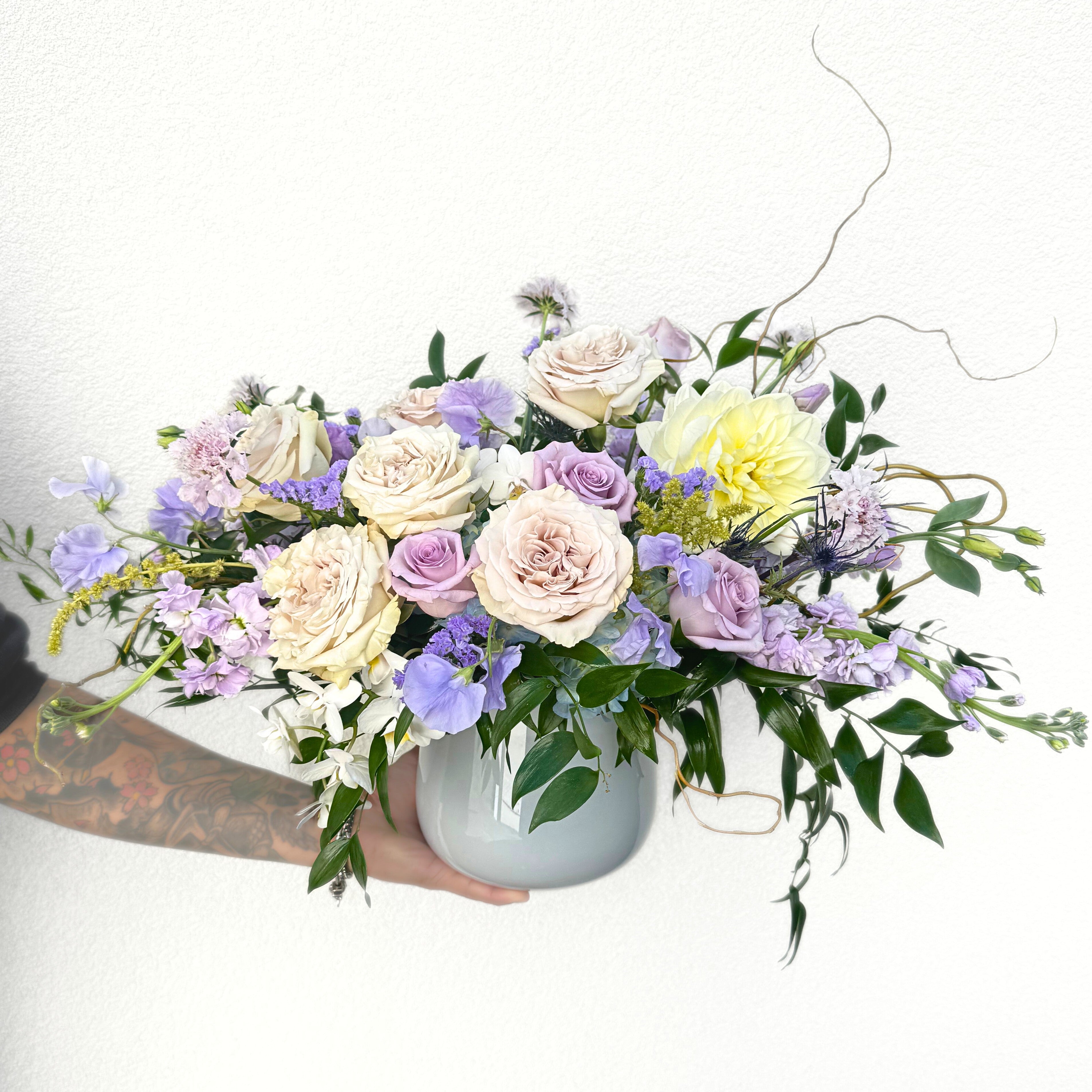 Lush mixed bouquet in white ceramic vase featuring cream roses, lavender and pink spray roses, purple lisianthus, yellow dahlia, and cascading greenery.