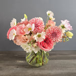 Pink and white dahlias, garden roses, and gerbera daisies arranged in a clear glass vase with lush green stems.