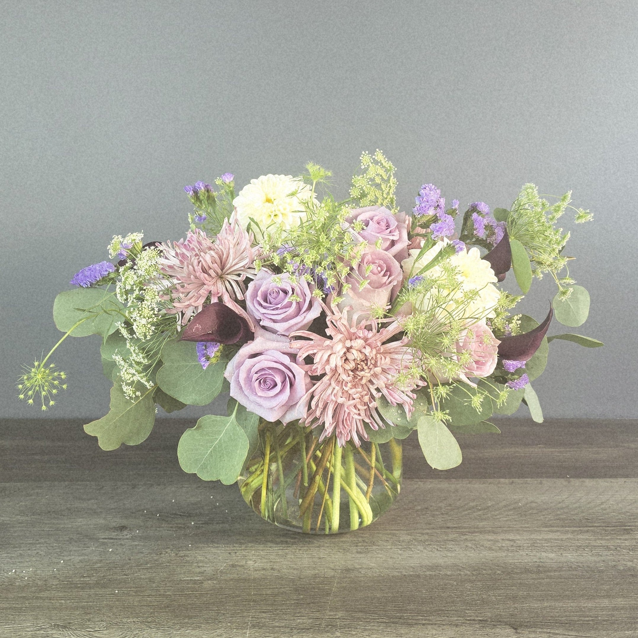 Lush glass vase arrangement with lavender roses, blush dahlias, white flowers, purple statice, and green hydrangea with eucalyptus foliage.