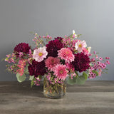 A lush bouquet of deep burgundy and pink dahlias, light pink gerbera daisies, and small purple flowers arranged in a clear glass vase with greenery.