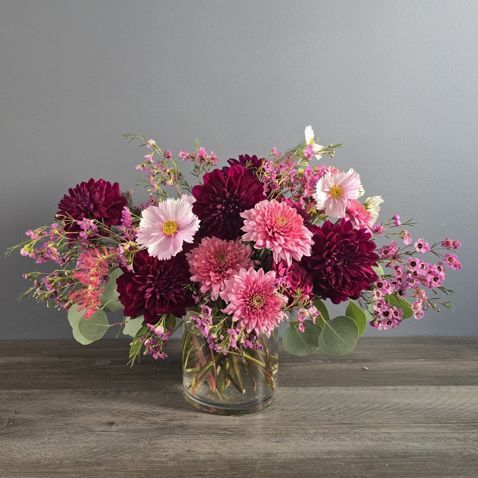 A lush bouquet of deep burgundy and pink dahlias, light pink gerbera daisies, and small purple flowers arranged in a clear glass vase with greenery.