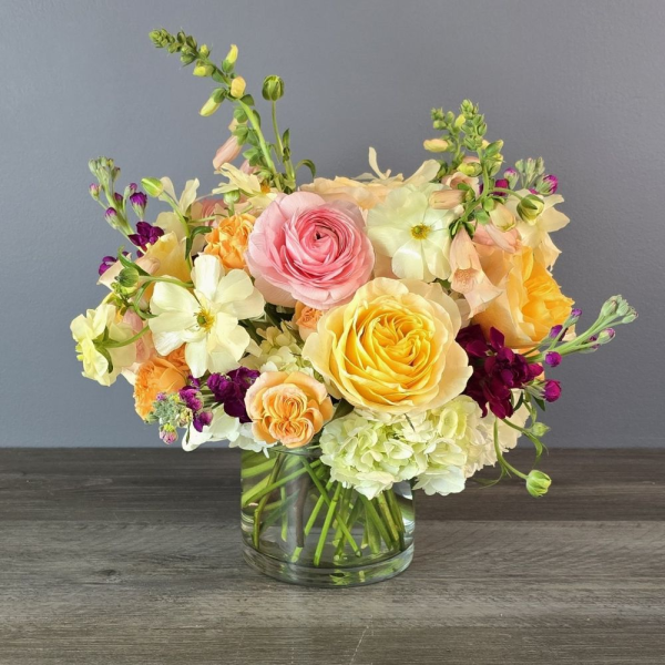 A lush mixed bouquet with pink and yellow roses, cream dahlias, coral ranunculus, deep burgundy spray roses, and green hypericum berries arranged in a clear…